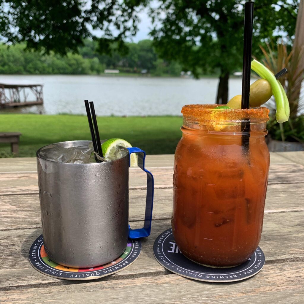 A mule in a metal mug and a Bloody Mary in a mason jar on an outdoor table, with Old Hickory Lake and a boat dock beyond the lawn.
